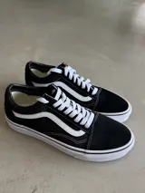 Championes Vans UA Old Skool, color negro con la icónica franja lateral Sidestripe™ blanca. Presentan una silueta baja con cordones blancos, parte superior de gamuza y lona resistentes, y cuello acolchado. La suela es de goma con patrón waffle.