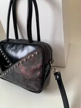 Cartera rígida negra con detalles en rojo y tachas plateadas. Tiene herrajes y cierre metálico plateado marca YKK, asa fina para llevar al hombro o en la mano y correa larga extra regulable. El interior es forrado, tiene un bolsillo con cierre enmarcado en cuero, y un segundo bolsillo plaqué.