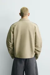 Sudadera de corte boxy fit color verde oliva, con cuello solapa y abertura frontal. Presenta mangas largas con bandas laterales negras a contraste y bordado frontal con texto en color negro.
