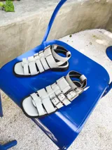 Sandalias planas de cuero color blanco hueso, con múltiples tiras horizontales y hebillas metálicas plateadas. Incluyen pulsera superior desmontable con hebillas laterales para regular el ajuste.