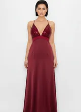 Vestido largo de fiesta color bordó, con escote en V profundo y estilo halter. El cuerpo superior presenta un diseño con lentejuelas brillantes, mientras que la falda es de corte fluido y liso.