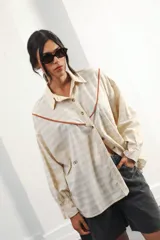 Camisa oversize de algodón color crema con rayas texturizadas, hombro caído y silueta amplia. Presenta un recorte delantero con vivo contrastante en color marrón, botones frontales y logo bordado en el frente.