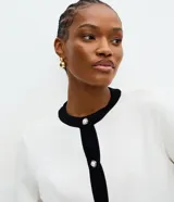 Cardigan de punto color blanco con detalles en negro en cuello, puños y tapeta frontal. Presenta cierre frontal con botones perlados y dos bolsillos delanteros falsos con solapa bicolor.