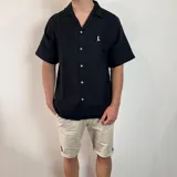 Camisa de hombre color verde oliva, de muselina con textura arrugada, manga corta, corte oversize, cuello cubano y cierre con botones. Presenta un pequeño logo bordado en el pecho.