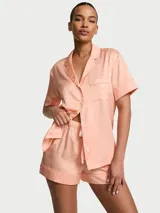 Pijama de satén color salmón, compuesto por una camisa de manga corta con botones frontales, cuello solapa y un bolsillo en el pecho, y un short con cintura ajustable.