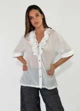 Blusa blanca de algodón con cuello en V, volados bordados en la pechera, cierre frontal con botones y mangas cortas con vuelta.