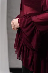 Blusa color bordó confeccionada en tul, con mangas largas y puños acampanados con volados, escote con lazo para atar en forma de corbata y volados superpuestos en el ruedo.