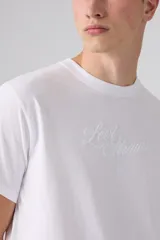 Remera blanca de manga corta y cuello redondo, con corte relajado y detalle de texto bordado en el pecho.