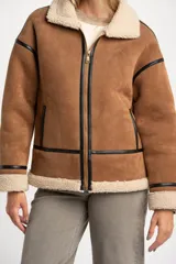 Campera tipo gamulán de corte oversize, confeccionada en material efecto gamuza color camel con interior y detalles de cuello y puños en corderito sintético color crema. Presenta cierre frontal y detalles de ribetes en cuero sintético negro.