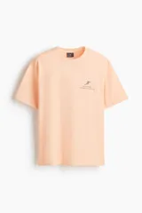 Remera color salmón de corte holgado, confeccionada en algodón, con cuello redondo y estampa pequeña en el pecho con la inscripción 'Echo Park Tennis Collective' y la silueta de un tenista.