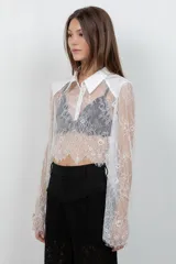 Camisa crop top de encaje blanco con mangas largas acampanadas, cuello camisero y pechera de tela lisa.
