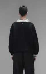 Sweater de punto negro con cuello polo en color crema, manga larga y diseño oversize. Presenta bordados en el frente con la inscripción 'Ravers', el número '90' y letras cursivas, con terminaciones en punto rib en puños y cuello.