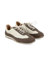 Zapatilla deportiva de mujer, estilo retro running, con combinación de colores blanco crudo y marrón oscuro. Presenta capellada de cuero y gamuza, cordones blancos y suela de goma color caramelo.