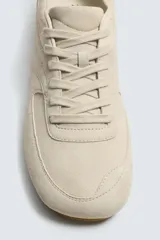 Zapatilla de cordones para hombre, confeccionada en piel con acabado de serraje en color beige. Presenta un diseño de perfil bajo con detalles de pespuntes decorativos y una suela ultrafina.