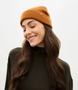 Gorro de punto color mostaza con borde plegado y pequeña etiqueta decorativa aplicada en el frente.