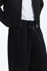 Jogger de punto de algodón color negro, con cintura elástica y cordón de ajuste, bolsillos laterales y pliegues frontales pespunteados. Corte relajado de pernera recta.