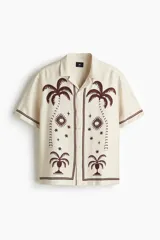 Camisa de manga corta color beige, con cuello abierto y cierre frontal de botones. Presenta un estampado en marrón de palmeras, soles y estrellas, con líneas en los bordes.