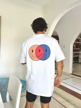 Remera blanca de corte oversize con estampado gráfico en la espalda que muestra dos círculos superpuestos formando un diagrama de Venn con caras expresivas en tonos naranja, rojo y azul.