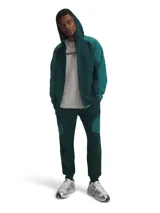 Pantalón de jogging Under Armour, modelo Unstoppable, en color verde oscuro. Presenta un diseño de doble punto con paneles en contraste, cintura elástica con cordón interno, bolsillos laterales abiertos y un bolsillo lateral con cierre. Corte ajustado en las piernas.