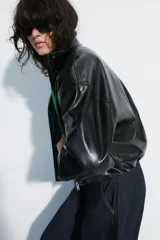 Chaqueta de cuero sintético negro con cuello alto, cierre frontal de cremallera oculto por solapa con botones a presión, mangas largas y bolsillos laterales. Diseño con volumen y bajo elástico.