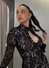 Vestido corto de encaje negro con forro en tono nude, de corte ajustado, cuello alto y abertura frontal tipo gota. Presenta mangas largas con terminación acampanada.