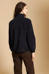 Camisa de pana color crema con cuello camisero con volados, cierre frontal de botones y mangas largas con puños abotonados. Presenta un bolsillo aplicado en el pecho y una silueta relajada.