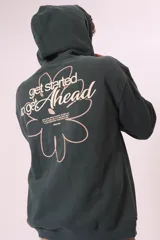 Buzo con capucha color verde oscuro, de corte holgado, con bolsillo frontal tipo canguro y cordones ajustables en el cuello. Presenta un pequeño bordado o estampado en el pecho con forma de flor y la frase 'get Ahead'.