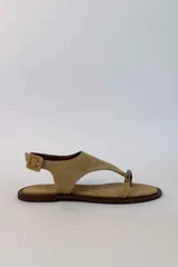 Sandalia plana de gamuza color tostado, con diseño de tira en T y argolla metálica en el empeine. Cuenta con tira ajustable con hebilla en el talón.