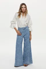 Camisa blanca de algodón con cuello alto, detalles de encaje broderie en el frente, mangas abullonadas y puños con volados y encaje. Cierre frontal con botones tapados.