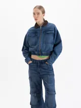 Campera bomber de denim azul con cierre frontal, cuello de pana en color marrón y bolsillos frontales con solapa. Presenta un diseño corto con cintura elástica.