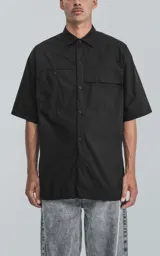 Camisa de manga corta en popelina de algodón, con diseño oversize y cierre frontal de botones. Presenta dos bolsillos delanteros, uno con solapa y otro con ojal y botón.