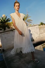Vestido largo color crudo, sin mangas, con escote en V pronunciado, canesú superior de tejido de punto con botones frontales y lazo para atar en el cuello, y falda fluida con detalle de bordado en el bajo.