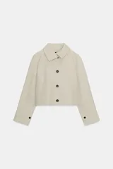Trench corta color beige con cuello solapa, manga larga con trabilla y botón en los puños, bolsillos de vivo en el delantero y cierre frontal con botones oscuros.