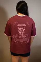 Remera oversize color bordó con cuello y puños de rib de color rosado. Estampado frontal en rosado con el texto "GUARDIÁN DE LA FUERZA" y otros símbolos.