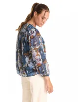 Blusa de gasa con estampado floral en tonos azules y marrones, con sutiles hilos de lurex dorados. Presenta cuello a la base, abertura central con botones, canesú delantero con pasamanería calada y mangas largas con puños elastizados. Incluye forro interno a tono.