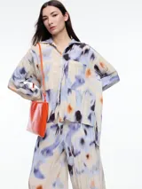 Camisa de manga larga confeccionada en viscosa crinkled con estampado de manchas abstractas en tonos azules, violetas, naranjas y negros sobre fondo beige. De silueta amplia y fluida, con bolsillos frontales grandes, tajos laterales y largo extendido en la espalda.