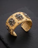 Brazalete rígido de bronce con baño dorado de la línea Imperial, diseño texturizado con apliques metálicos ornamentales en tono plateado, incrustaciones de circonias y piedras negras ovales centrales.