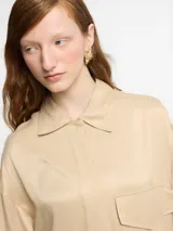Camisa color beige de tejido satinado, corte oversize, manga larga, tapeta con botones invisibles y logo bordado en la espalda.