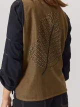 Chaleco de paño con diseño bicolor, frente en color negro y espalda en color marrón con textura de espiga. Presenta un maxi bordado de hoja en la parte posterior y detalles de tejido en los bordes.