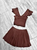 Conjunto color marrón chocolate, compuesto por un top corto con escote en V, breteles finos, volados en los hombros y elástico en la cintura, y una minifalda plisada con cintura alta.