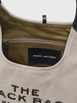 Cartera hobo Marc Jacobs The Belted Sack de lona color beige claro, con detalles en cuero marrón y el texto "THE SACK BAG MARC JACOBS" estampado en el frente.