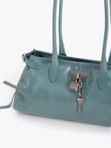 Cartera de hombro confeccionada en cuero vacuno color azul petróleo con acabado brillante. Presenta un diseño rectangular con herraje metálico tipo candado y dijes colgantes en el frente, además de correas laterales ajustables con tiras decorativas.