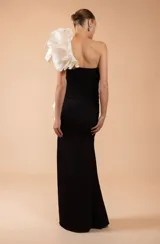 Vestido largo de fiesta color negro, con diseño ajustado y fruncido en el torso. Presenta un escote asimétrico con un gran aplique de volados estructurados en color blanco que recorre el hombro y el lateral de la falda, la cual cuenta con un tajo profundo.