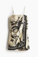 Vestido corto strapless con frente cruzado y diseño drapeado. Presenta un estampado tropical en tonos beige y negro con motivos de flamencos y palmeras. Incluye breteles finos ajustables opcionales y cierre lateral oculto.