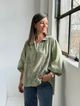 Camisa verde claro de corte oversize, con mangas tres cuartos abullonadas, escote en V con cuello camisero y cierre frontal parcial de botones. Cuenta con delicados bordados florales tono sobre tono distribuidos por toda la prenda.