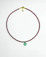 Collar corto compuesto por pequeñas cuentas facetadas de color bordó y un dije colgante circular de piedra verde con detalle dorado en el centro.