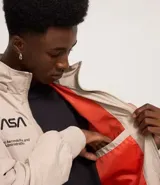 Campera rompevientos color beige con capucha, cierre frontal y bolsillos laterales. Presenta estampado de la NASA en el pecho, parche circular en la manga derecha y bandera de Estados Unidos en la manga izquierda.