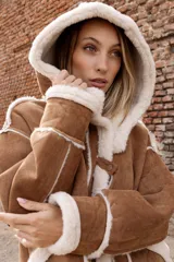 Campera estilo gamulán color tostado con interior y bordes de corderito sintético color crema. Presenta capucha amplia, cierre frontal con trabas y broches metálicos, y bolsillos delanteros grandes. Posee un calce relajado y diseño de textura suave.