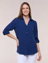 Blusa de calce holgado y caída fluida color azul, con cuello camisero, escote en V y cierre parcial de tres botones metálicos. Tiene mangas largas convertibles a tres cuartos mediante presilla, textura sutilmente arrugada tipo crepé y ruedo curvo.