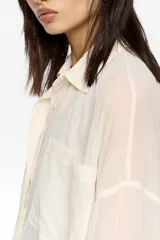 Camisa larga de corte oversize color crema, confeccionada en tejido plisado. Presenta cuello camisero, botones frontales y bolsillo plastrón en el pecho.
